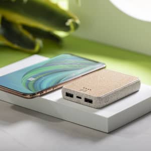A power bank in a beige finish sits beside a smartphone on a white platform, with greenery in the background.