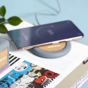 A wireless charger in grey with a cork centre, featuring a smartphone placed on top, resting on a colourful magazine page.
