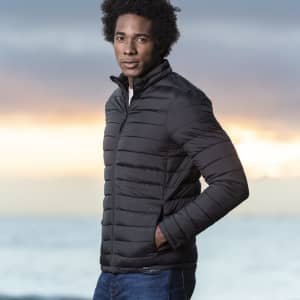 A man wearing a black puffer jacket, standing against a scenic background of water and sunset.