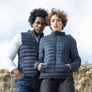 A navy puffer jacket worn by a man and a woman, featuring a zip front and side pockets. The image has a natural outdoor backdrop.