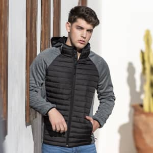 A soft shell jacket in black and grey, featuring a zip-up front and a hood, displayed against a sunlit wall.