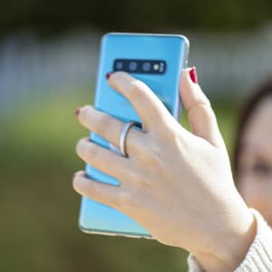 A hand holding a blue smartphone with a ring holder, set against a blurred outdoor background.