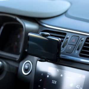 A car phone holder in black attached to a vehicle's dashboard air vent. The dashboard features various controls and displays.