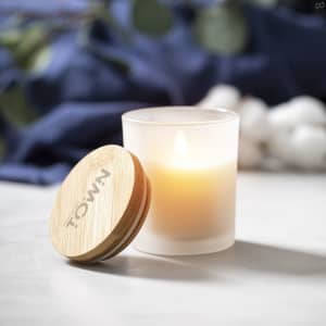 A frosted glass candle with a bamboo lid, featuring a soft, warm light, set against a blue backdrop and cotton plants.
