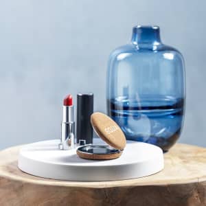 A round pocket mirror in a cork case placed on a white surface, alongside a red lipstick and a blue glass vase.
