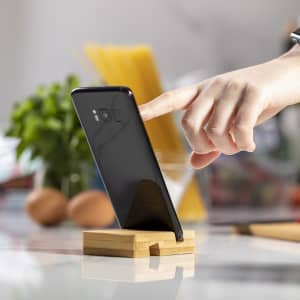 A black smartphone resting on a wooden phone stand, with a hand pointing at the screen and various kitchen items in the background.