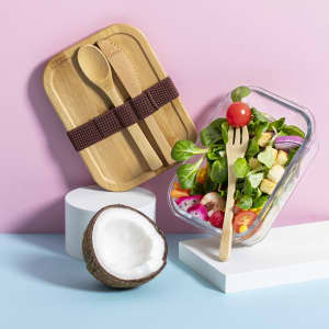 A bamboo lunch box with utensils sits beside a glass container filled with a fresh salad and a coconut on display.