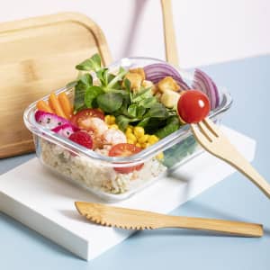 A glass container filled with a salad featuring shrimp, tomatoes, corn, and various vegetables, with wooden cutlery beside it.