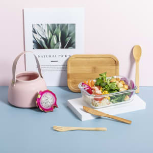 A pink lunch box beside a wooden cutting board, bamboo utensils, a drink bottle, and a dragon fruit on a pastel background.