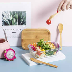 A glass lunch box filled with salad and vegetables, accompanied by a bamboo serving spoon and wooden fork.