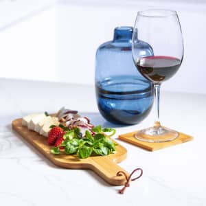 A bamboo chopping board with cheese, strawberries, and greens, alongside a wine glass and a blue drink bottle on a coaster.