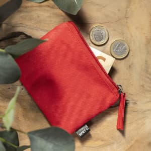 A red zippered purse sits on a wooden surface, accompanied by two coins and surrounded by greenery.