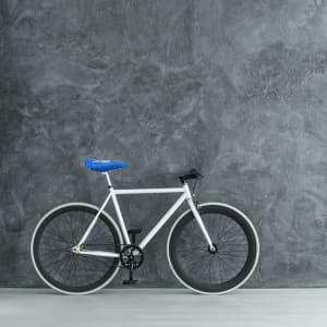 A white bicycle with a blue saddle cover against a textured grey wall, featuring black wheels and green accents.