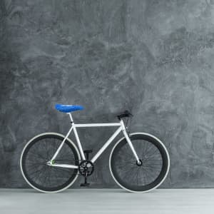 A bicycle with a white frame, black wheels, and a blue saddle cover, positioned against a textured grey wall.