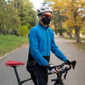 A cyclist wearing a blue jacket and black pants stands next to a bicycle with a red saddle cover in a park.