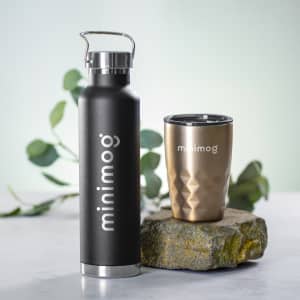 A drink bottle in black beside a gold insulated cup, both featuring a logo, set on a stone surface.