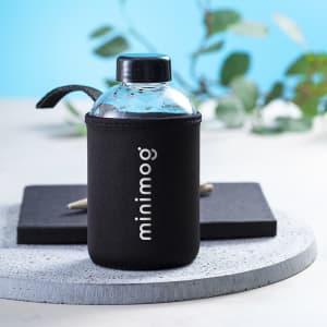 A drink bottle with a clear body and a black sleeve, featuring a black lid and a logo. Set on a grey circular surface.