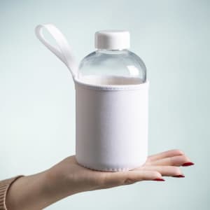 A drink bottle with a transparent body and white sleeve, held in a hand. It features a white cap and a carrying handle.