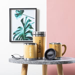 A set of bamboo insulated cups in various sizes with stainless steel details, displayed on a table beside a plant print.