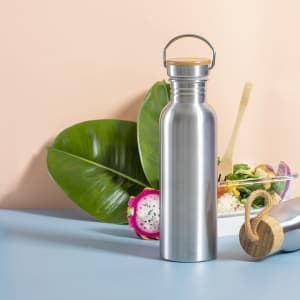 A drink bottle in stainless steel with a wooden lid, placed among tropical leaves and a fruit backdrop.