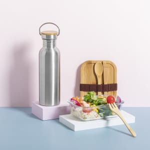 A drink bottle in stainless steel with a wooden lid, beside a salad in a glass container and wooden utensils.