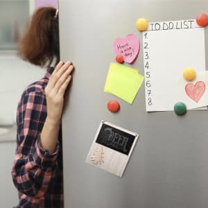A grey fridge covered with colourful magnets and notes, with a person partially hidden behind it in a checked shirt.