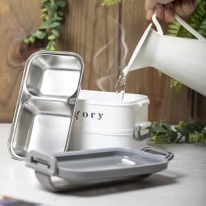 A grey thermal lunch box with a stainless steel insert and a white container is shown, accompanied by a pouring jug.