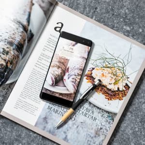 A black and gold Zabox Stylus Touch Ball Pen positioned on a magazine page with food photography and a smartphone.