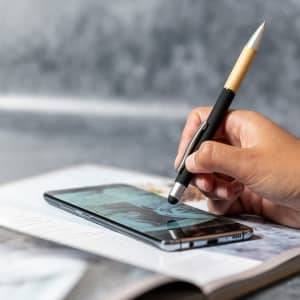 A black stylus touch ball pen with a silver clip is being used on a smartphone screen, placed on a magazine.