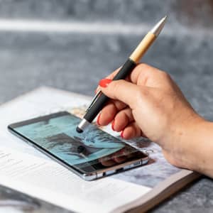 A black and bamboo stylus touch ball pen held above a smartphone on a magazine page. Features a logo on the pen.