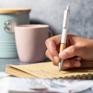 A metal pen with a white and wooden design rests above a notebook, while a pastel mug and container are in the background.