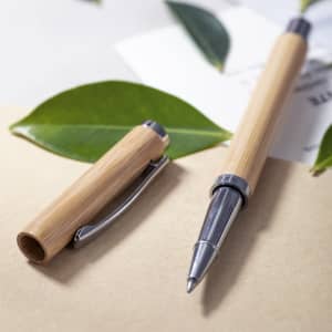 A bamboo pen with a silver clip and nib, placed beside its cap on a textured surface with green leaves.