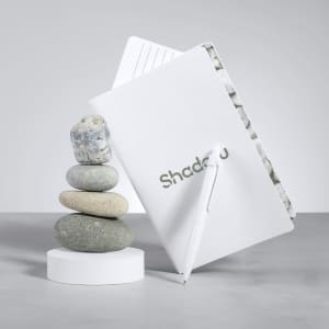 A white notebook with a patterned interior and a white pen resting beside a stack of smooth stones on a light surface.