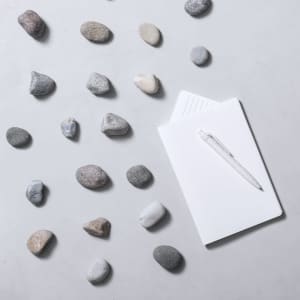A clear pen rests on a white notebook, surrounded by smooth grey and beige stones on a light surface.