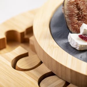 A bamboo cheese board with a slate centre, featuring sliced cheese and a fig. The board has cut-out cheese shapes.