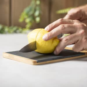 A bamboo cutting board with a black surface, featuring a hand slicing a yellow lemon with a knife.