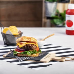 A black chopping board with a burger topped with cheddar, lettuce, tomato, and red onion, set against a striped backdrop.
