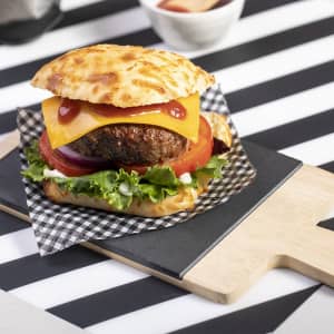 A fresh burger with cheese, lettuce, tomato, and onion sits on a black cutting board, surrounded by a checkered paper liner.
