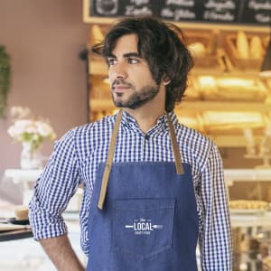 A denim apron with a logo is worn by a man with dark hair in a café setting. He is dressed in a checked shirt.