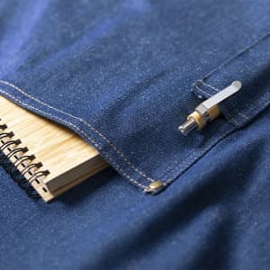 A denim apron featuring a pocket with a small notepad and a pen, plus a branded logo.