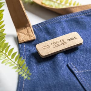 A denim apron with a tan strap featuring a wooden name tag and a branding logo. Green leaves are in the background.