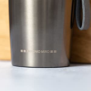 A stainless steel vacuum bottle with a grey strap and a branding logo near the base.