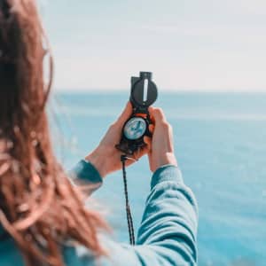 A person holding a black compass with a strap, overlooking a blue sea under a clear sky.