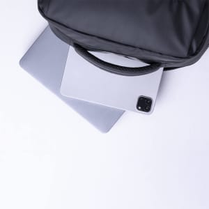 A black backpack has a handle and is placed over a light grey laptop and a tablet. It features a logo.