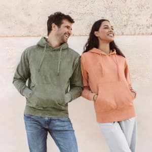 Two people wearing hoodies: one in olive green and the other in peach, both with front pockets and hoods.