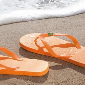 Bright orange flip flops resting on sandy beach, with a logo visible. Waves gently lapping at the shore nearby.