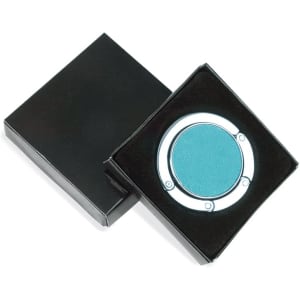 A turquoise foldable bag hanger inside a black presentation box, featuring a simple design and a metallic rim.
