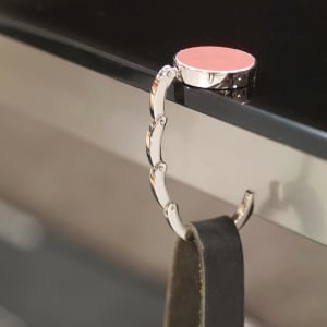 A foldable bag hanger in silver metal with a pink top, designed to clip onto tables or surfaces.