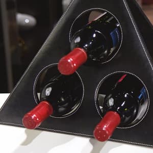 A drink bottle holder featuring a triangular design with three circular openings for red wine bottles. The holder is black with white stitching.