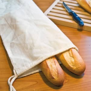 A drawstring bread bag in natural fabric holds two fresh baguettes, with a knife and cutting board in the background.